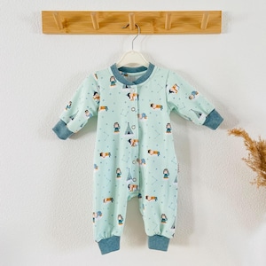 Könnte beinhalten: Ein hellblauer Baby-Strampler mit einem Muster aus kleinen Figuren und Zelten. Der Strampler hat lange Ärmel und Druckknöpfe auf der Vorderseite.