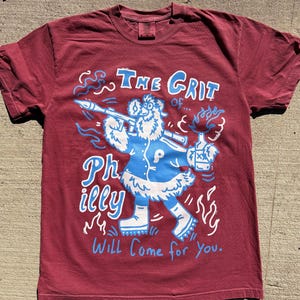 May include: A maroon t-shirt with a graphic design. The design features a cartoon character in a blue coat and white boots holding a rocket launcher and a bottle. Text on the shirt reads "The Grit of... Philly Will Come for You."