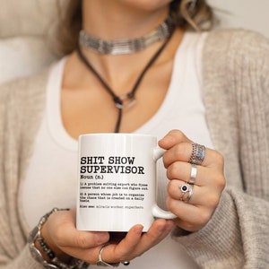 May include: White ceramic coffee mug with a black and white design that reads "Shit Show Supervisor" and a definition of the term. The mug is being held by a person's hand.