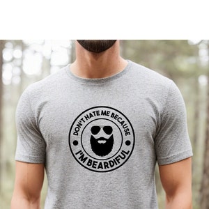 May include: A grey t-shirt with a circular graphic that reads "DON'T HATE ME BECAUSE I'M BEARDIFUL." The graphic features a beard, sunglasses, and text in a black circle. The shirt is short-sleeved.