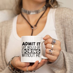 May include: White ceramic mug with the text "ADMIT IT LIFE WOULD BE BORING WITHOUT ME" in black. The mug has a handle and is held by a person wearing rings and bracelets.