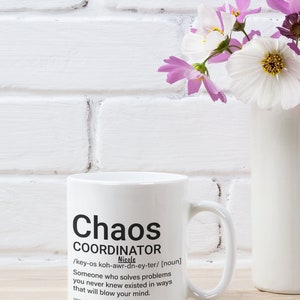 May include: White ceramic mug with a black design that reads "Chaos Coordinator" and a definition of the term. The mug has a black handle and is sitting on a wooden surface.
