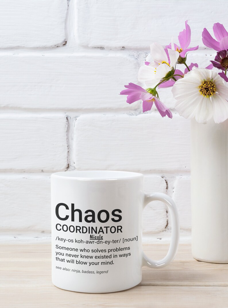 May include: White ceramic mug with a black design that reads "Chaos Coordinator" and a definition of the term. The mug has a black handle and is sitting on a wooden surface.
