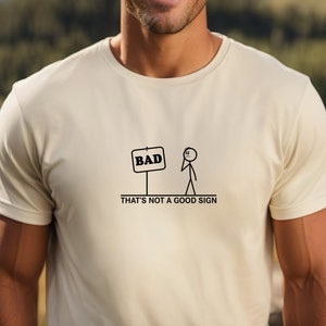May include: A white t-shirt featuring a stick figure illustration and the text "BAD" on a sign. The text below the illustration reads "THAT'S NOT A GOOD SIGN."