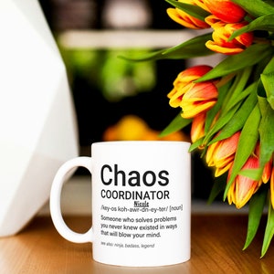 May include: White ceramic mug with a black graphic that reads "Chaos Coordinator" and a definition of the term. The mug is sitting on a wooden table with a bouquet of orange and yellow tulips in the background.