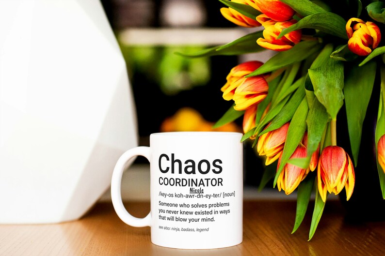 May include: White ceramic mug with a black graphic that reads "Chaos Coordinator" and a definition of the term. The mug is sitting on a wooden table with a bouquet of orange and yellow tulips in the background.