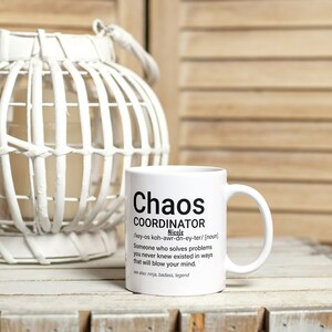 May include: White ceramic mug with black text that reads "Chaos Coordinator" and a definition of the word. The mug is sitting on a wooden surface next to a white wicker lantern.