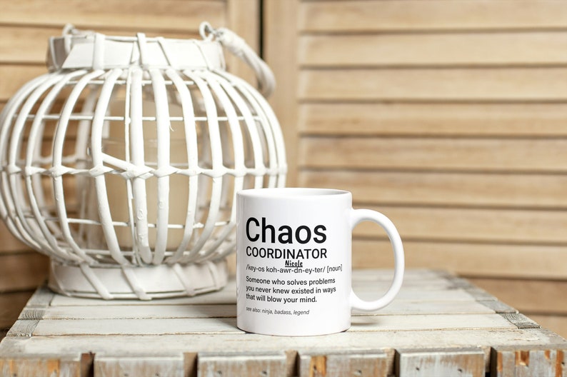 May include: White ceramic mug with black text that reads "Chaos Coordinator" and a definition of the word. The mug is sitting on a wooden surface next to a white wicker lantern.
