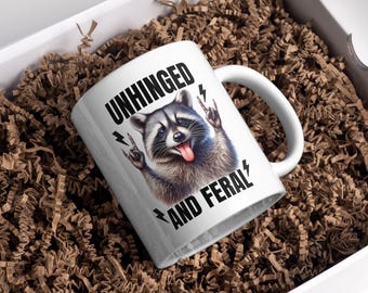 May include: White ceramic mug featuring a raccoon illustration. The raccoon is sticking out its tongue and making a rock-and-roll hand gesture. The words "UNHINGED AND FERAL" are printed on the mug with lightning bolt graphics.