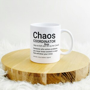 May include: White ceramic mug with a black graphic that reads "Chaos Coordinator" and a definition of the word. The mug is sitting on a wooden coaster.