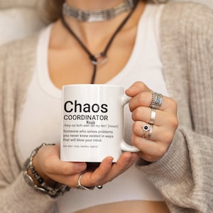 May include: White ceramic mug with a black and white definition of "Chaos Coordinator" with the name Nicole. The mug is being held by a person wearing a white shirt and a brown sweater.