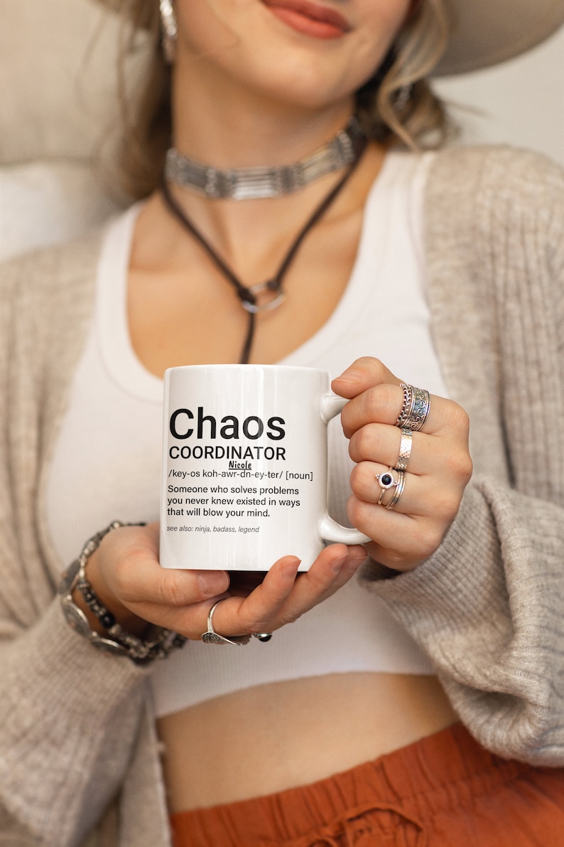 May include: White ceramic mug with a black and white definition of "Chaos Coordinator" with the name Nicole. The mug is being held by a person wearing a white shirt and a brown sweater.