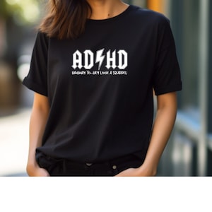 Może przedstawiać: Czarny t-shirt z białym napisem "ADHD" nad "HIGHWAY TO...HEY LOOK A SQUIRREL" w stylizowanej czcionce. Koszulka ma krótkie rękawy i okrągły dekolt.