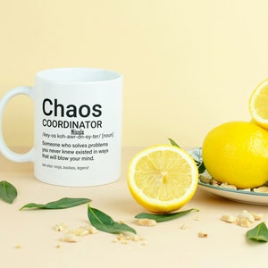 May include: White ceramic mug with a black design that reads "Chaos Coordinator" and a definition of the word. The mug is sitting on a light brown surface with lemons and green leaves.