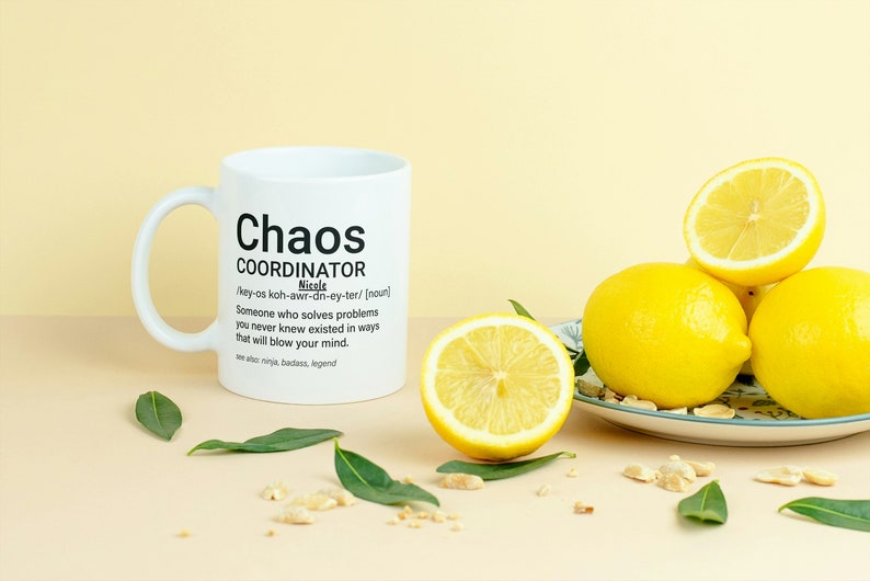 May include: White ceramic mug with a black design that reads "Chaos Coordinator" and a definition of the word. The mug is sitting on a light brown surface with lemons and green leaves.