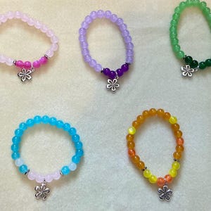 May include: Five beaded bracelets in various colors: light pink, purple, green, blue, and orange. Each bracelet features a flower charm. The bracelets are made of small, round beads.