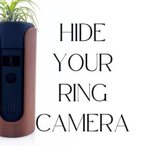 Könnte beinhalten: Ein brauner und schwarzer Pflanzer mit einer kleinen Luftpflanze darin. Der Pflanzer hat einen Schlitz für eine Ring-Kamera, und der Text "HIDE YOUR RING CAMERA" ist in Schwarz auf weißem Hintergrund geschrieben.