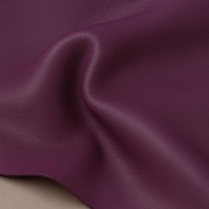 May include: Close-up of a piece of deep purple leather. The leather is folded and textured, with subtle variations in color and tone. The material appears soft and supple, with a slight sheen. The background is a neutral beige.