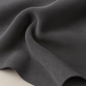 May include: Close-up of a dark gray fabric with a textured surface. The fabric is draped, creating soft folds and shadows. The material appears to be smooth and flowing, suggesting a lightweight textile.