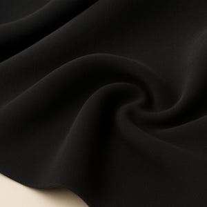 May include: Close-up of a black fabric with soft folds and a smooth texture. The fabric appears to be flowing, with subtle highlights and shadows that emphasize its drape and quality. The overall impression is of a luxurious material.
