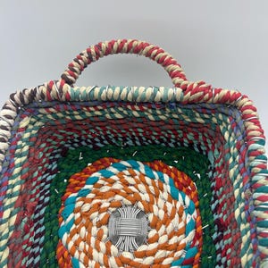 Puede incluir: Una cesta tejida multicolor con forma cuadrada y asa. La cesta presenta una vibrante mezcla de hilos rojos, verdes, azules y naranjas. El interior tiene un diseño circular en el centro. El asa es un diseño de cuerda trenzada.