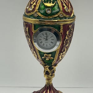 May include: An ornate, egg-shaped clock with a red, green, and gold color scheme. The clock features a small, round face with Roman numerals and is embellished with jewels and floral designs. The clock is approximately 6 inches tall.