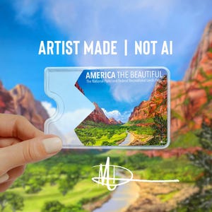 May include: A clear plastic America the Beautiful National Parks Pass with a scenic landscape image. The pass is held against a backdrop of a blue sky and red rock mountains. Text on the pass reads "ARTIST MADE | NOT AI".