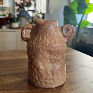 Op de afbeelding: Een kleine, getextureerde keramische vaas in tinten perzik en crème. De vaas heeft een cilindrisch lichaam met twee lusvormige handvatten en een kleine opening aan de bovenkant. Het oppervlak is bedekt met kleine, verhoogde stippen.
