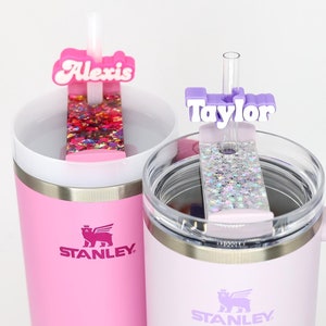 May include: Two Stanley tumblers, one pink and one lavender, each with a personalized straw topper. The pink tumbler has a topper that says "Alexis" with colorful glitter. The lavender tumbler has a topper that says "Taylor" with silver glitter.