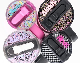May include: Four circular tumbler lids with sliding closures, arranged in a cluster. Two lids feature the name Alexis in pink or white lettering. The designs include colorful confetti, black and pink glitter, and a black and white checkerboard pattern.