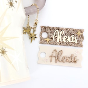 May include: A cream-colored tumbler with a gold starburst design, paired with two name tags that read "Alexis." One tag is glittery gold, the other is cream-colored. A gold charm chain with star and animal charms is attached.