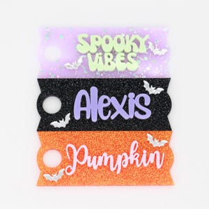 May include: Stacked Halloween-themed tags in purple, black, and orange glitter. The top tag reads "Spooky Vibes" in green, the middle tag says "Alexis" in purple, and the bottom tag says "Pumpkin" in white. Silver bat accents are scattered throughout.