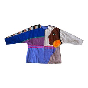 May include: A long-sleeve jacket featuring a color-blocked design with panels of blue, purple, and light gray. The right side displays a large applique of a face with a brown complexion, green eye, red lips, and a large orange circular earring.