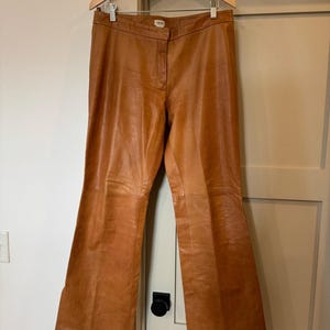 May include: A pair of tan leather trousers featuring a mid-rise waistband, a front zip fly with a button fastening, and a straight-leg cut. The trousers are displayed on a wooden hanger, showcasing the smooth texture and consistent tan colour throughout.