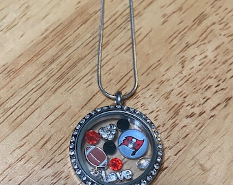 Puede incluir: Un collar de plata con un dije en forma de medallón con un inserto de vidrio transparente. El medallón contiene una pequeña pelota de fútbol, un corazón, una cuenta negra, un strass rojo y un logotipo de los Tampa Bay Buccaneers. La palabra "love" está escrita en letras plateadas.