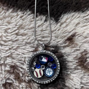 May include: A silver-toned necklace with a round, clear pendant filled with baseball-themed charms. The charms include a baseball, a baseball logo, and a compass. The pendant is surrounded by small, clear crystals and the word "love".