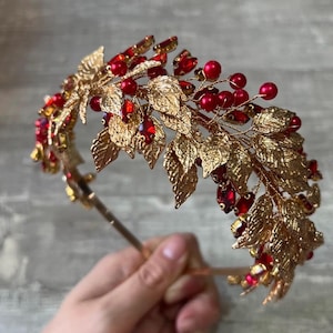 Könnte beinhalten: Ein goldfarbener Metallkranz mit roten Beeren und roten Schmuckakzenten. Der Kranz hat goldfarbene Blätter und ein dünnes Band. Der Kranz ist für das Tragen auf dem Kopf konzipiert, wahrscheinlich für einen besonderen Anlass.