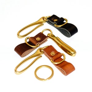 May include: Three brass keychains with leather loops in brown, tan, and black. The keychains have a hook and a ring.