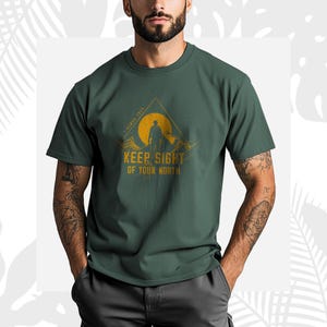 May include: Dark green t-shirt with a mountain and sun graphic, and the text "KEEP SIGHT OF YOUR NORTH". The design features a person's silhouette. The graphic is in yellow and orange tones.