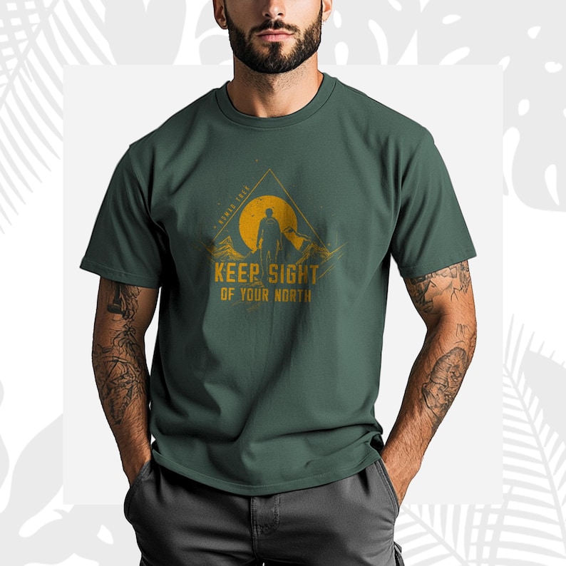 May include: Dark green t-shirt with a mountain and sun graphic, and the text "KEEP SIGHT OF YOUR NORTH". The design features a person's silhouette. The graphic is in yellow and orange tones.