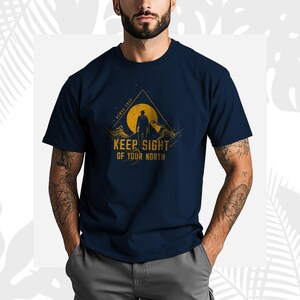 May include: Navy blue t-shirt with a graphic design featuring a silhouette of a person on a mountain, a yellow sun, and the text "KEEP SIGHT OF YOUR NORTH". The shirt has short sleeves.