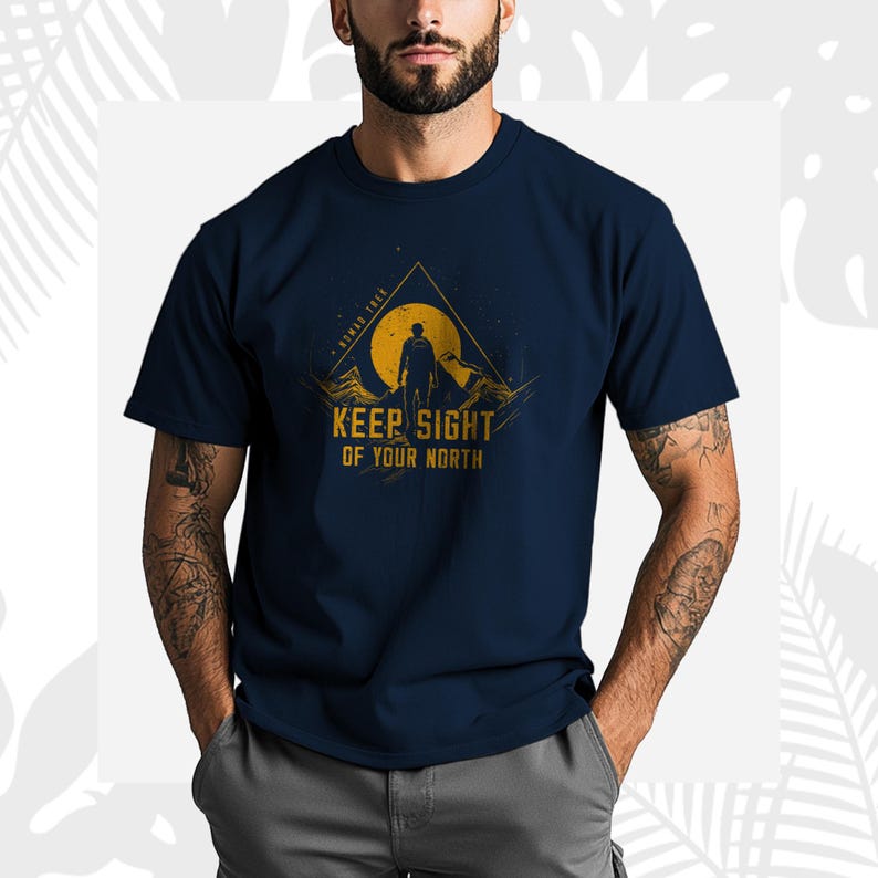 May include: Navy blue t-shirt with a graphic design featuring a silhouette of a person on a mountain, a yellow sun, and the text "KEEP SIGHT OF YOUR NORTH". The shirt has short sleeves.