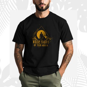 May include: Black t-shirt with a graphic design of a person standing before a mountain and a sun. The text "KEEP SIGHT OF YOUR NORTH" is printed below the graphic in a bold, sans-serif font. The design has a yellow and orange color scheme.
