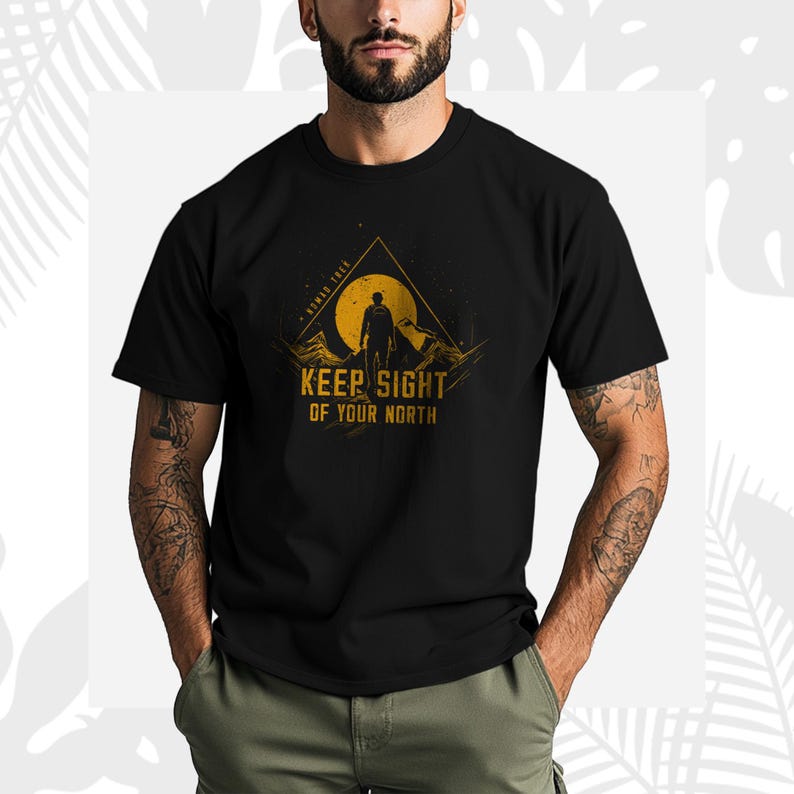 May include: Black t-shirt with a graphic design of a person standing before a mountain and a sun. The text "KEEP SIGHT OF YOUR NORTH" is printed below the graphic in a bold, sans-serif font. The design has a yellow and orange color scheme.