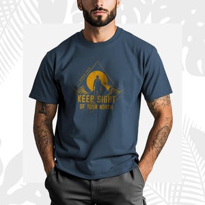May include: Navy blue t-shirt with a mountain and sun graphic and the text "KEEP SIGHT OF YOUR NORTH". The design includes a silhouette of a person. The shirt is made of a soft material.