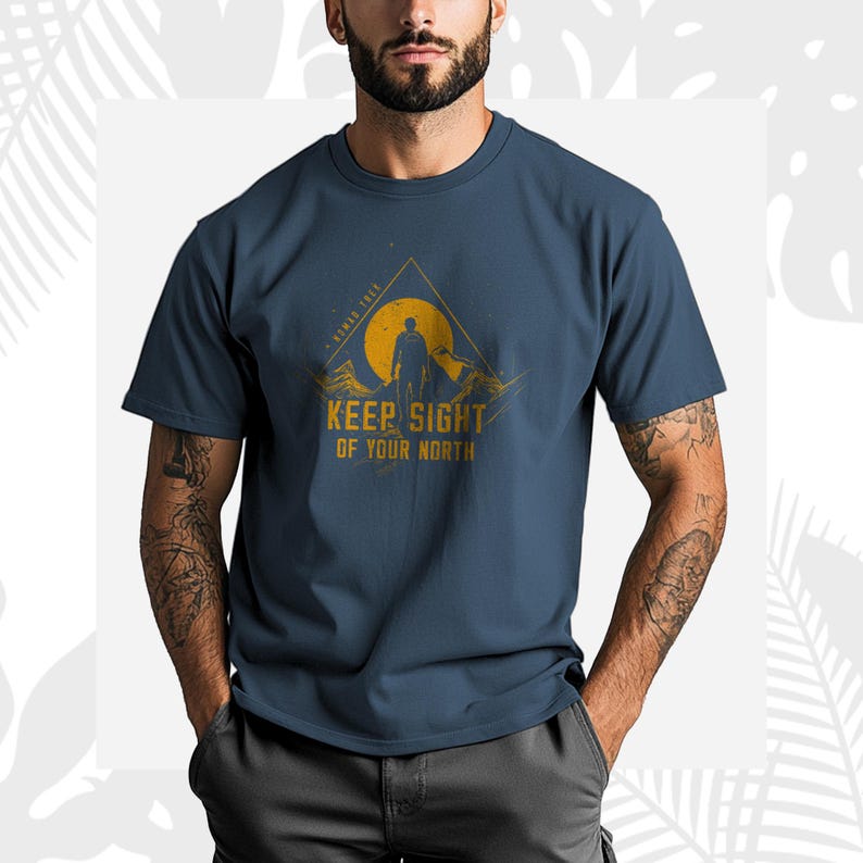 May include: Navy blue t-shirt with a mountain and sun graphic and the text "KEEP SIGHT OF YOUR NORTH". The design includes a silhouette of a person. The shirt is made of a soft material.