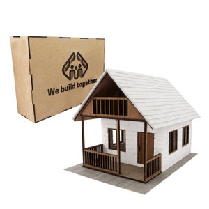 Puede incluir: Una casa modelo de madera blanca y marrón con un techo blanco y un pequeño porche. La casa está sentada sobre una superficie gris. La caja en el fondo dice "We build together".
