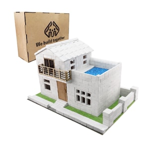 Puede incluir: Un modelo de casa de bloques de espuma blanca con un balcón, una piscina en el techo y una pequeña valla. La casa tiene una puerta de madera marrón y una barandilla de madera marrón en el balcón. La casa está sentada sobre una base verde con una acera blanca. La caja en el fondo tiene el texto "We build together" y un logotipo de dos personas dándose la mano.