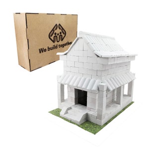 May include: A white building made of toy bricks with a tiled roof and a small porch. The building is sitting on a green base. The box in the background says "We build together".
