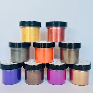 May include: Eight small jars of mica powder in various colors, including gold, copper, purple, and brown. The jars are arranged in a pyramid shape on a white surface.